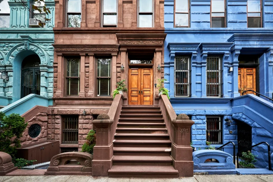 Colorful teal, brown, and blue brownstones in New York City with wide stone stairways going up to first floor doors.